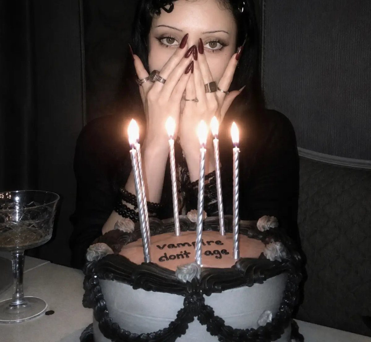 Woman in goth attire posing behind a grey cake with lit candles.