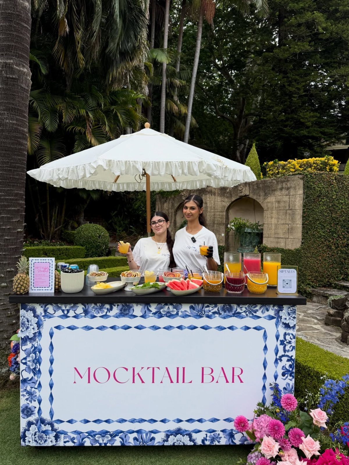 Mocktail bar with blue floral patterns, colorful drinks, and fruit in a garden.