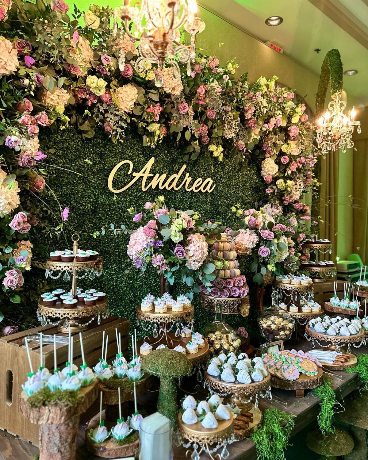 Dessert table with gold stands and sweets before a floral greenery wall.