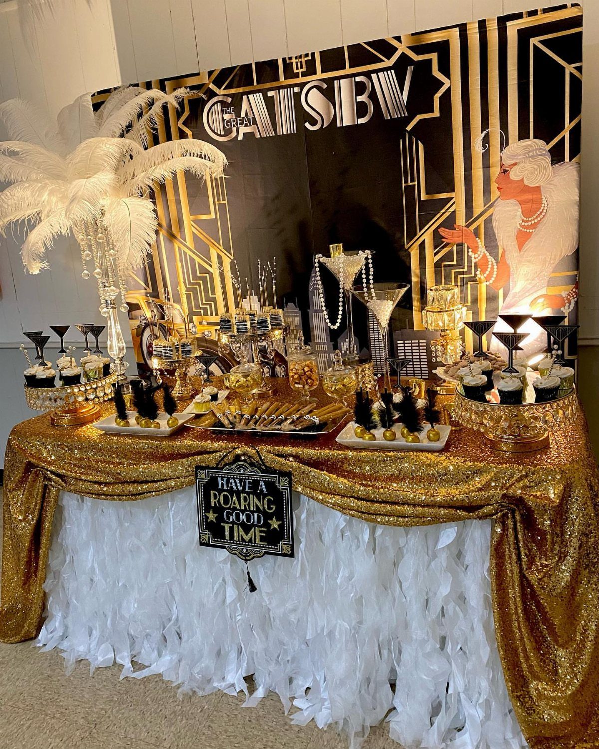 Gold and white Gatsby themed party table with sequins, feathers, and cupcakes.