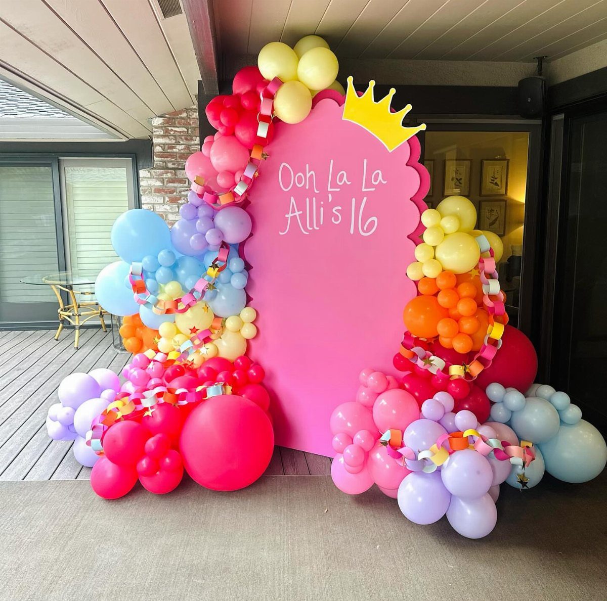 Pink backdrop with a yellow crown and colorful balloon arch for a sixteenth birthday.