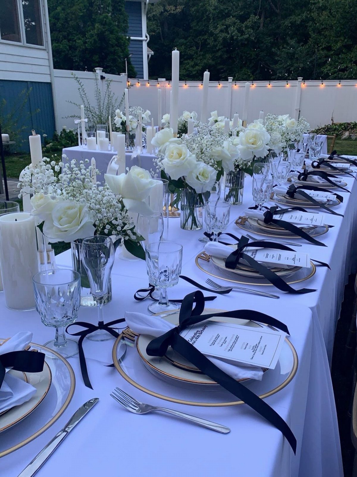 Elegant outdoor table setting with white roses, gold plates, and black ribbons.