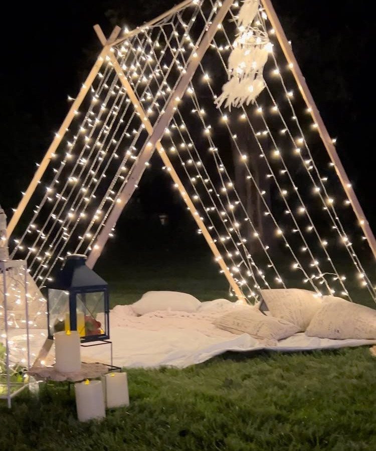 Wooden A-frame tent draped in fairy lights with pillows and blankets on grass