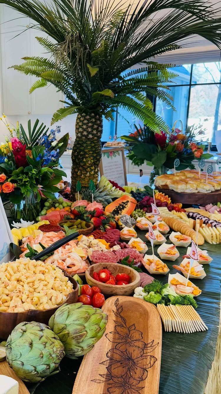 Tropical food spread with a pineapple palm tree centerpiece and colorful floral arrangements.