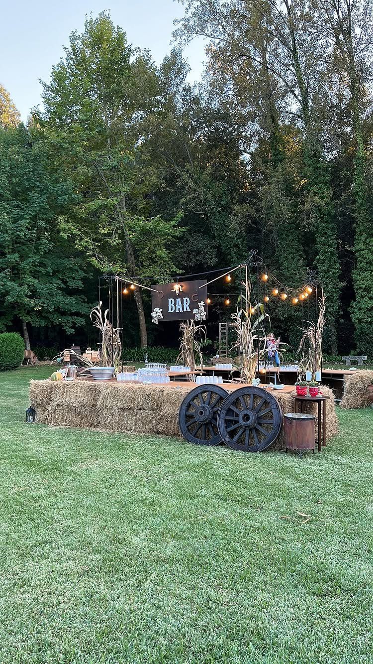 Rustic outdoor bar with hay bales, wagon wheels, and corn stalks on grass.