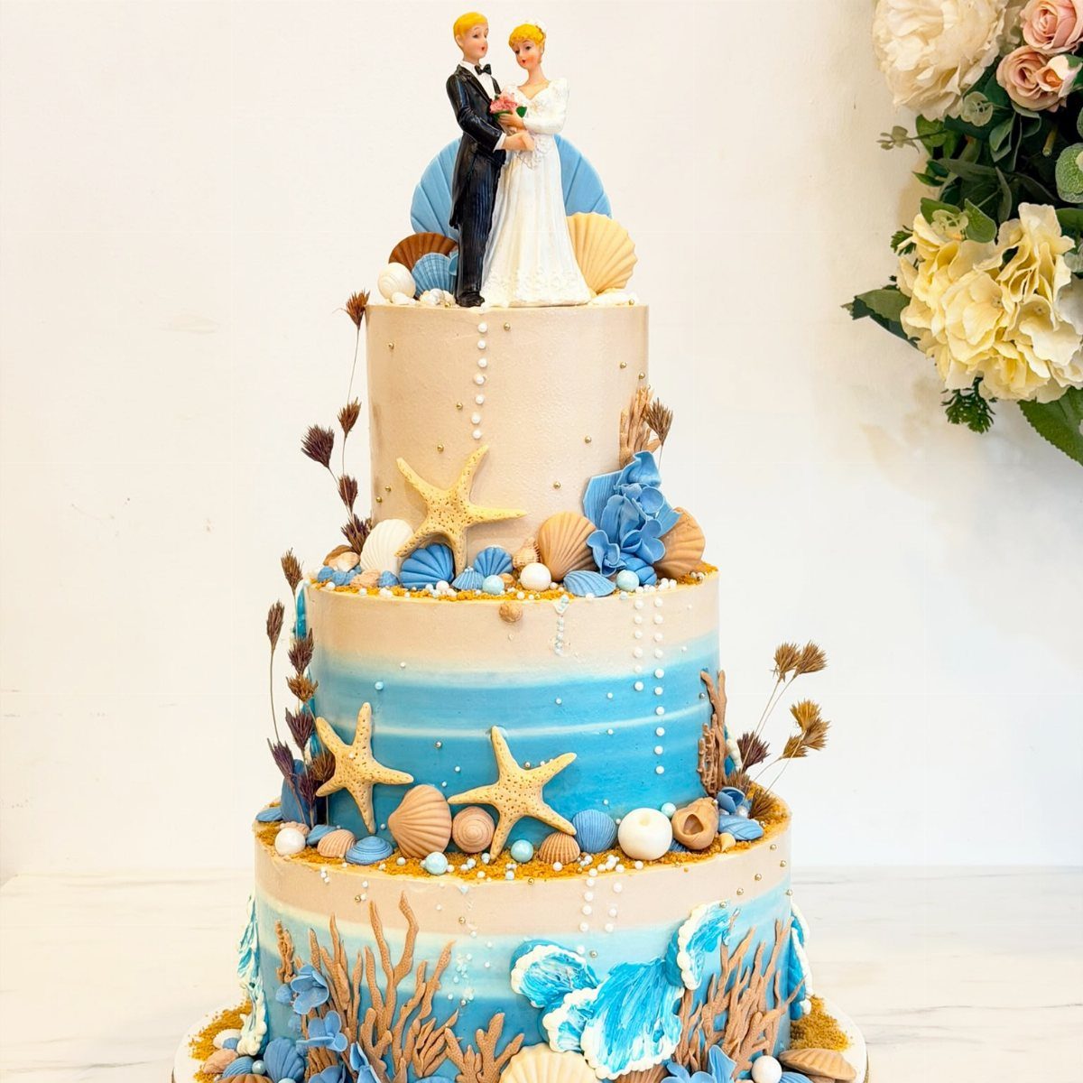 Three-tier beach themed cake with blue frosting, seashells, starfish, and wedding couple topper.