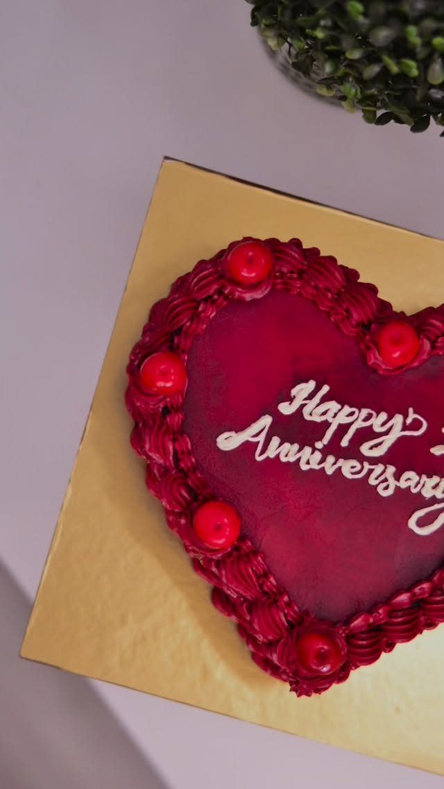 Heart-shaped red anniversary cake with cherries and white icing text on gold board
