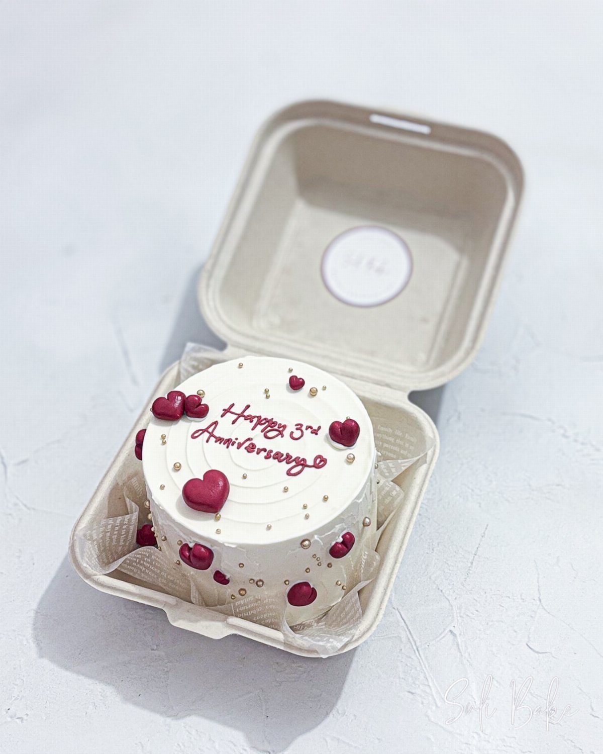 Small white anniversary bento cake with red hearts and cursive lettering in container