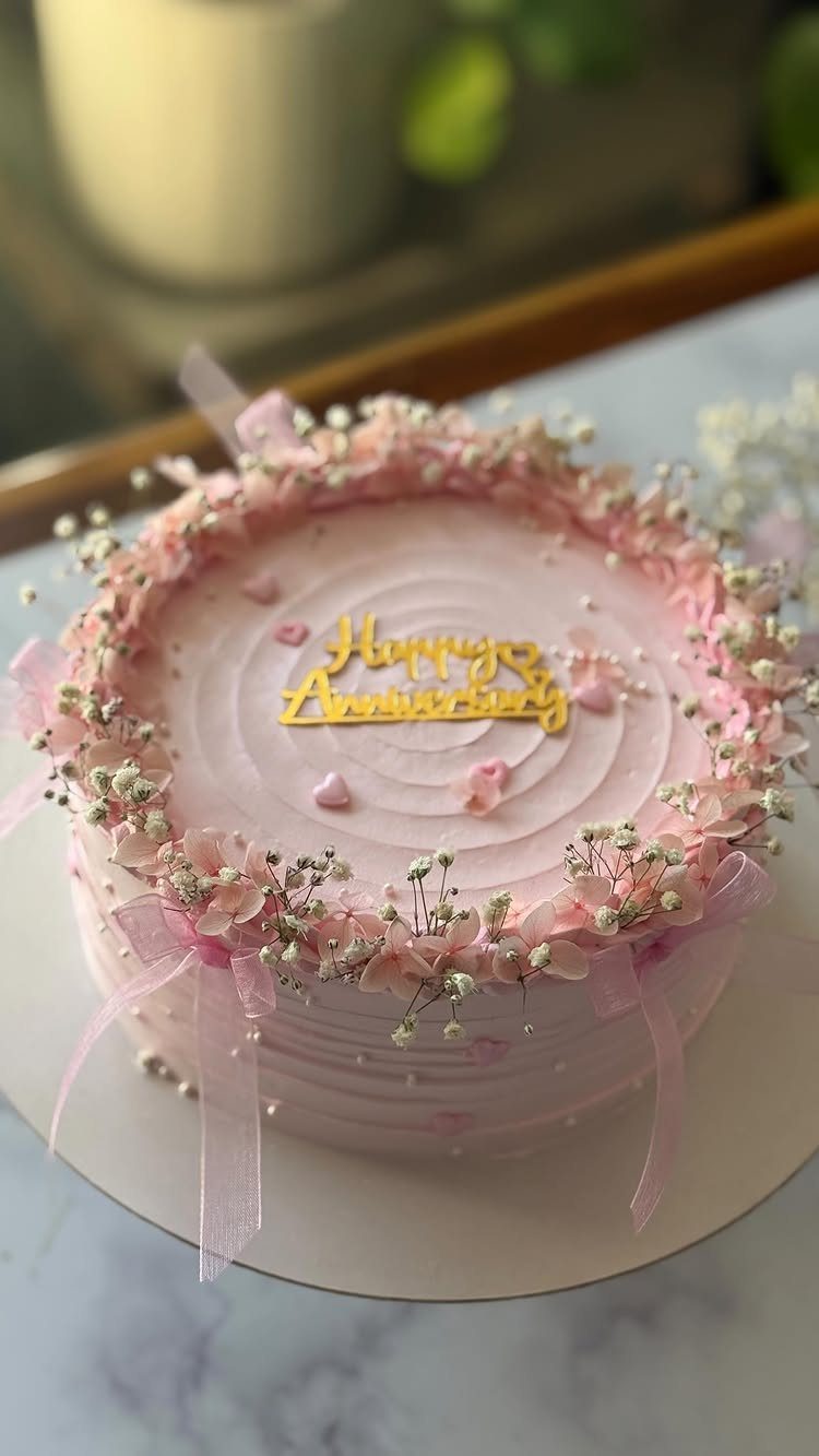 Pink anniversary cake decorated with delicate pink flowers, ribbons, and a gold topper.
