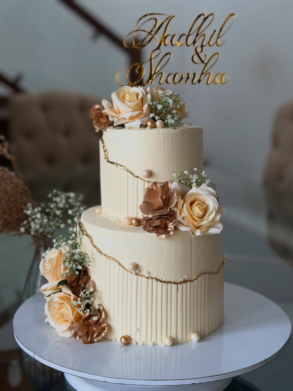 Two-tier white cake with gold glitter accents, cream roses, and a gold name topper.