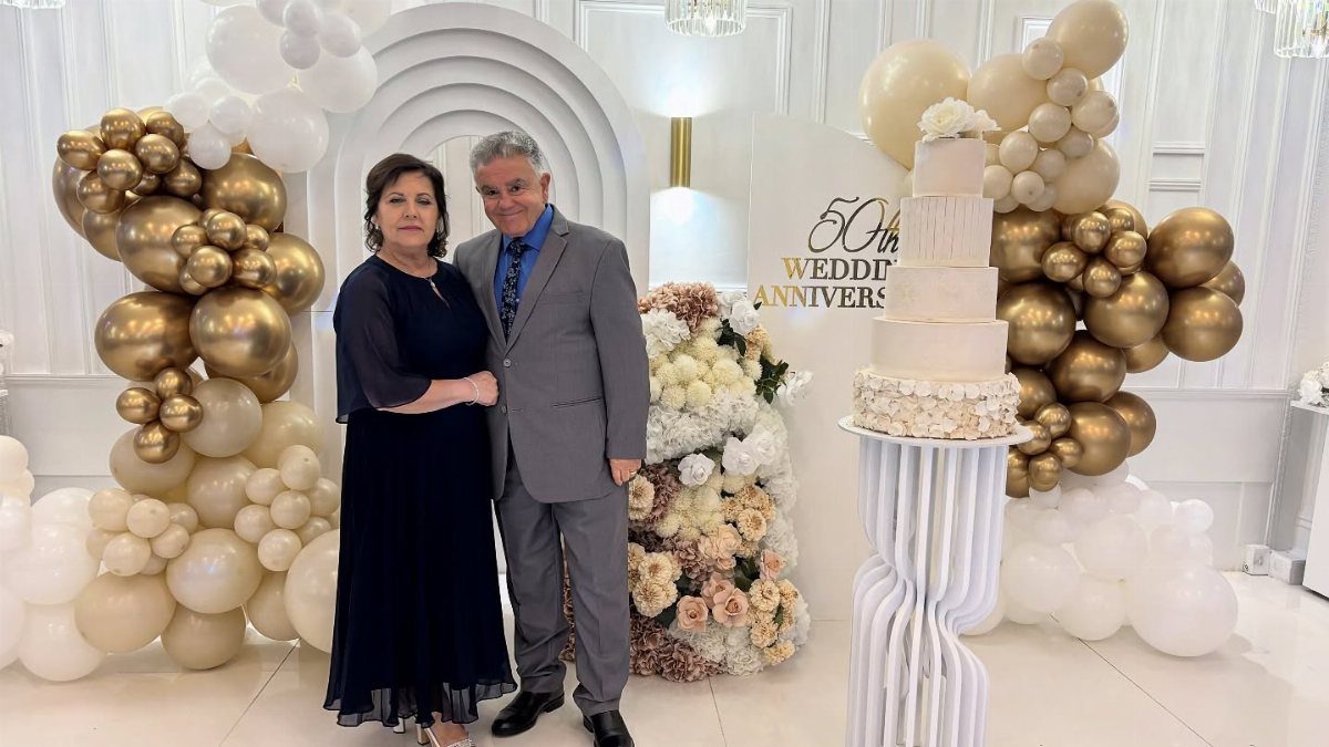 Couple posing at their 50th wedding anniversary party with balloons and a cake.