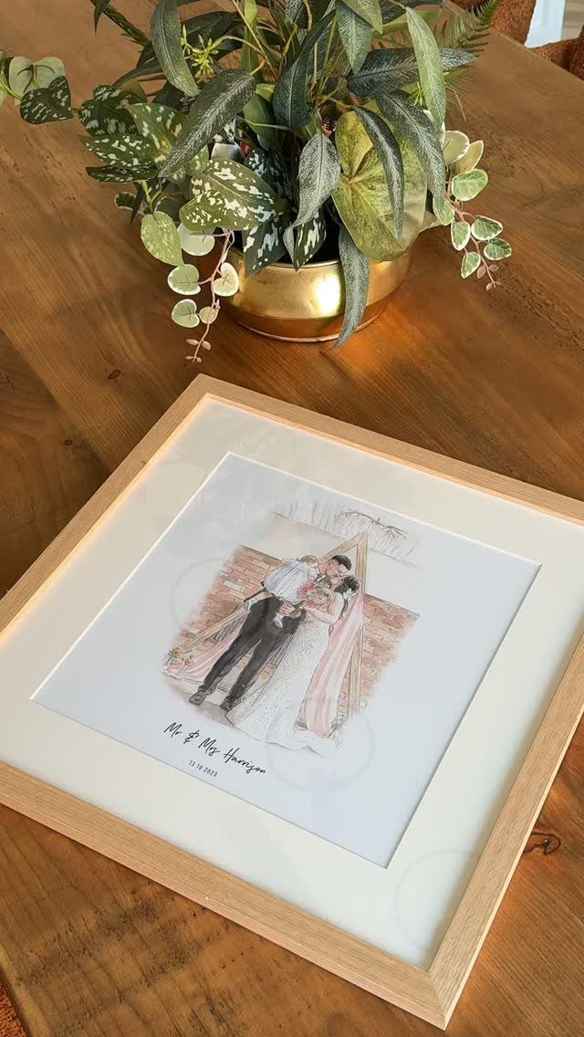 Framed couple illustration on a wooden table next to a potted green plant