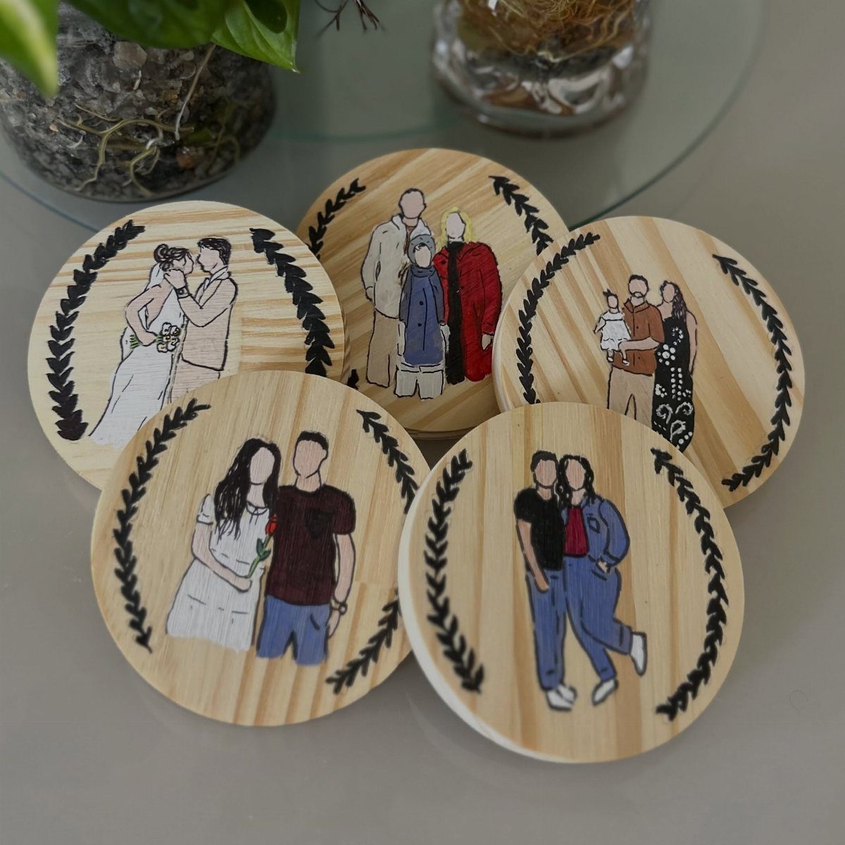 Five wooden coasters with custom family illustrations framed by black laurel wreaths.