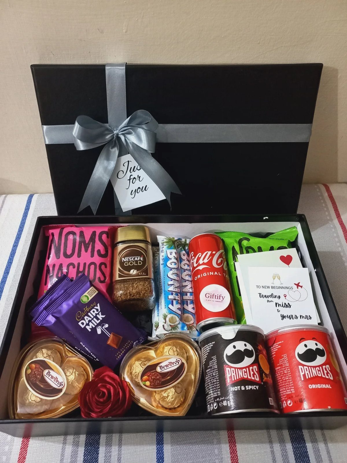 Black gift box filled with assorted snacks, chocolates, and drinks with a silver ribbon.