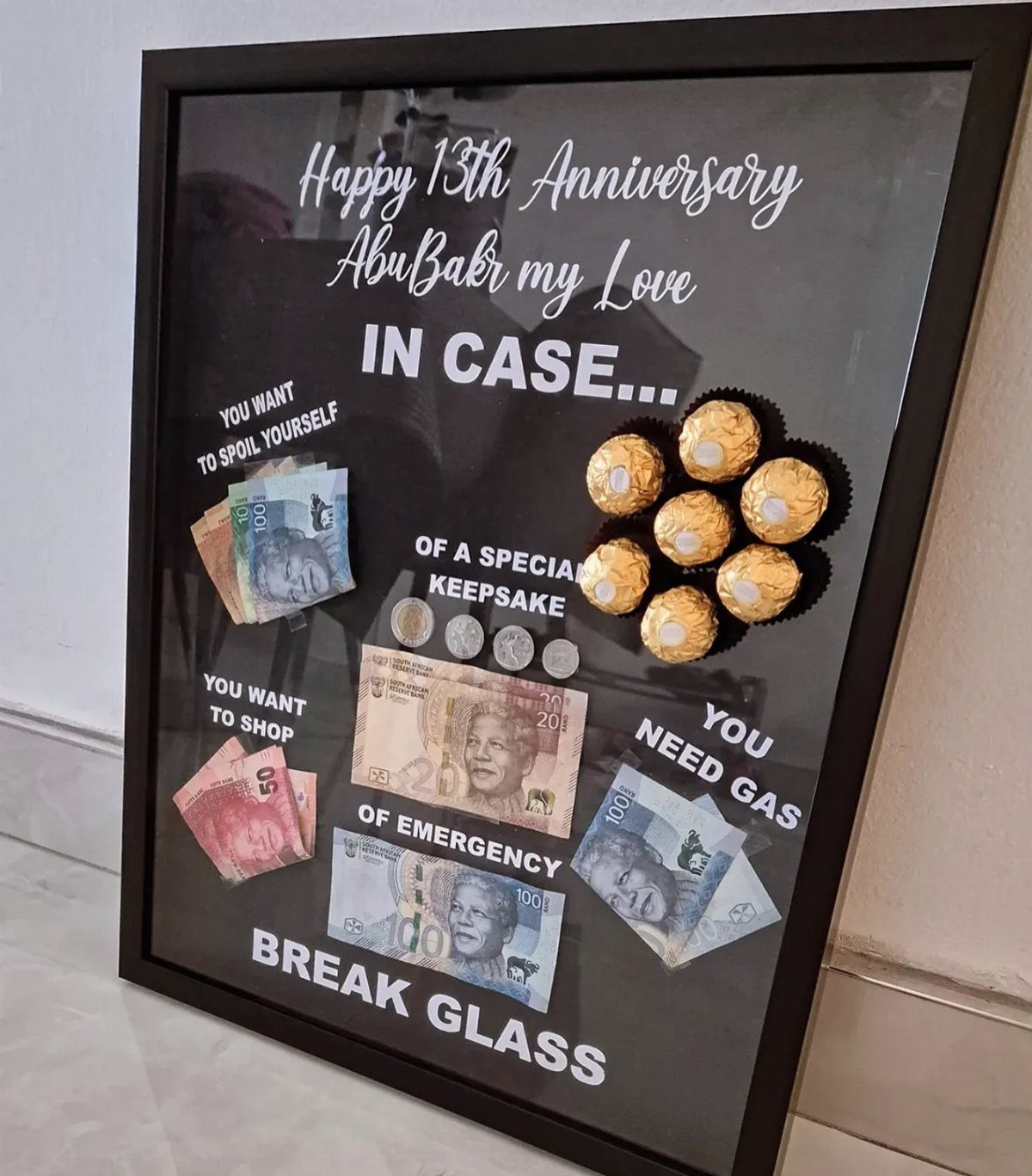 Framed anniversary gift box with cash, coins, and chocolates behind glass.